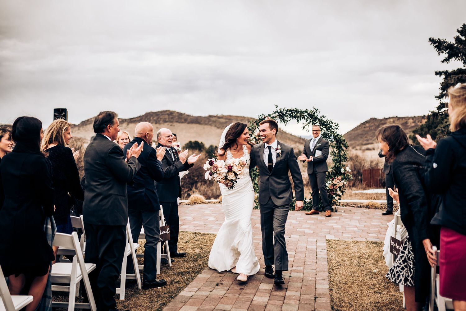 Newlyweds walk down stone aisle while wedding guests applaud against mountain backdrop.