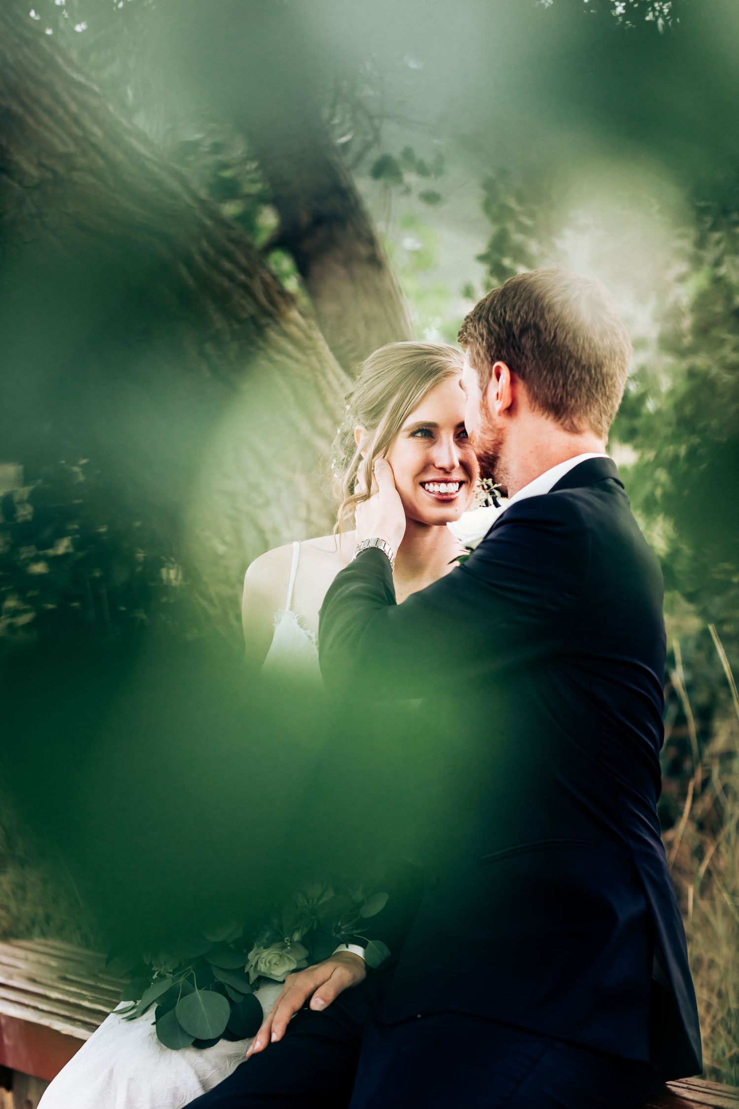 Intimate moment between newlyweds captured through green foliage in garden setting.