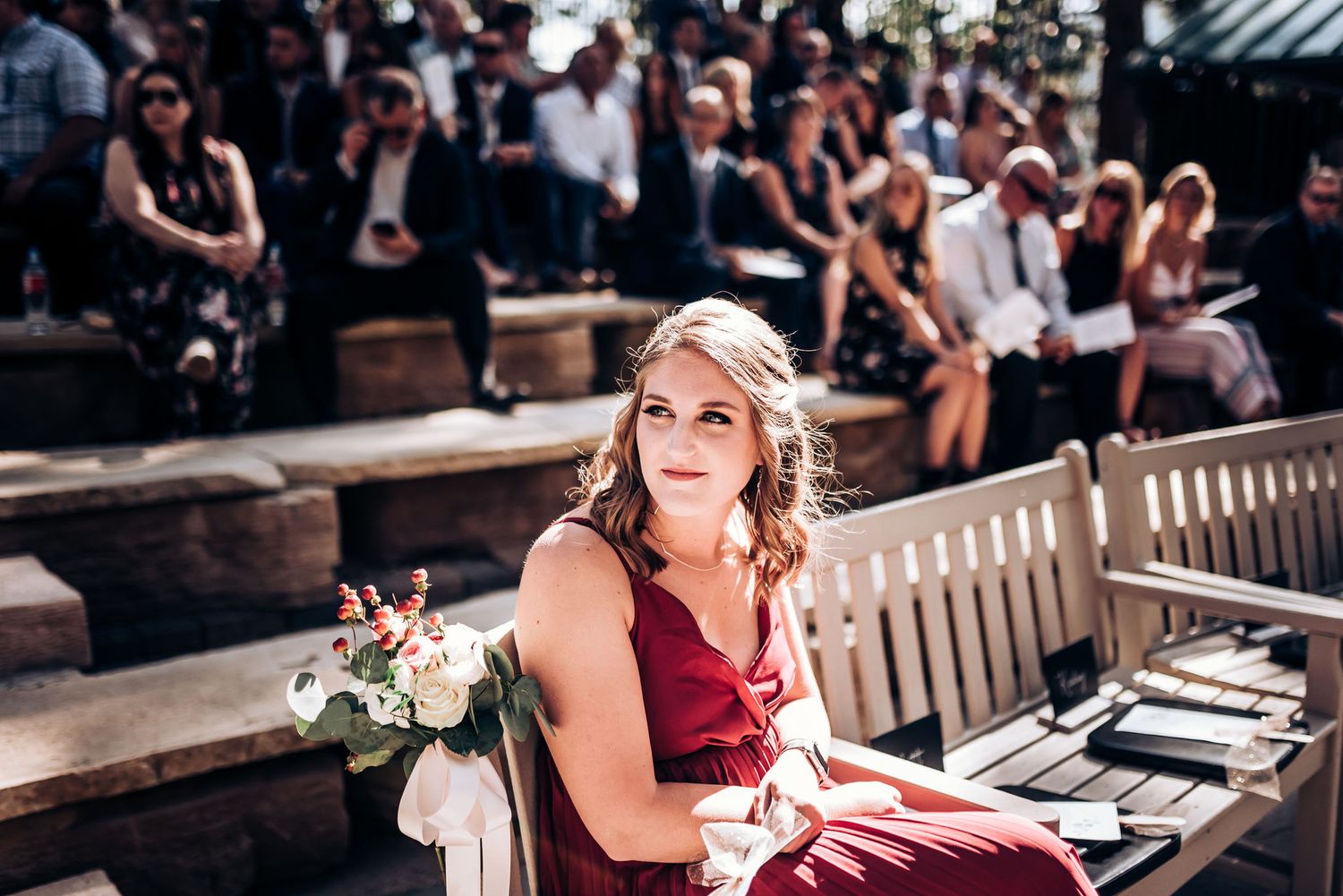 A bridesmaid in burgundy dress sits on wooden benches at an outdoor wedding venue.
