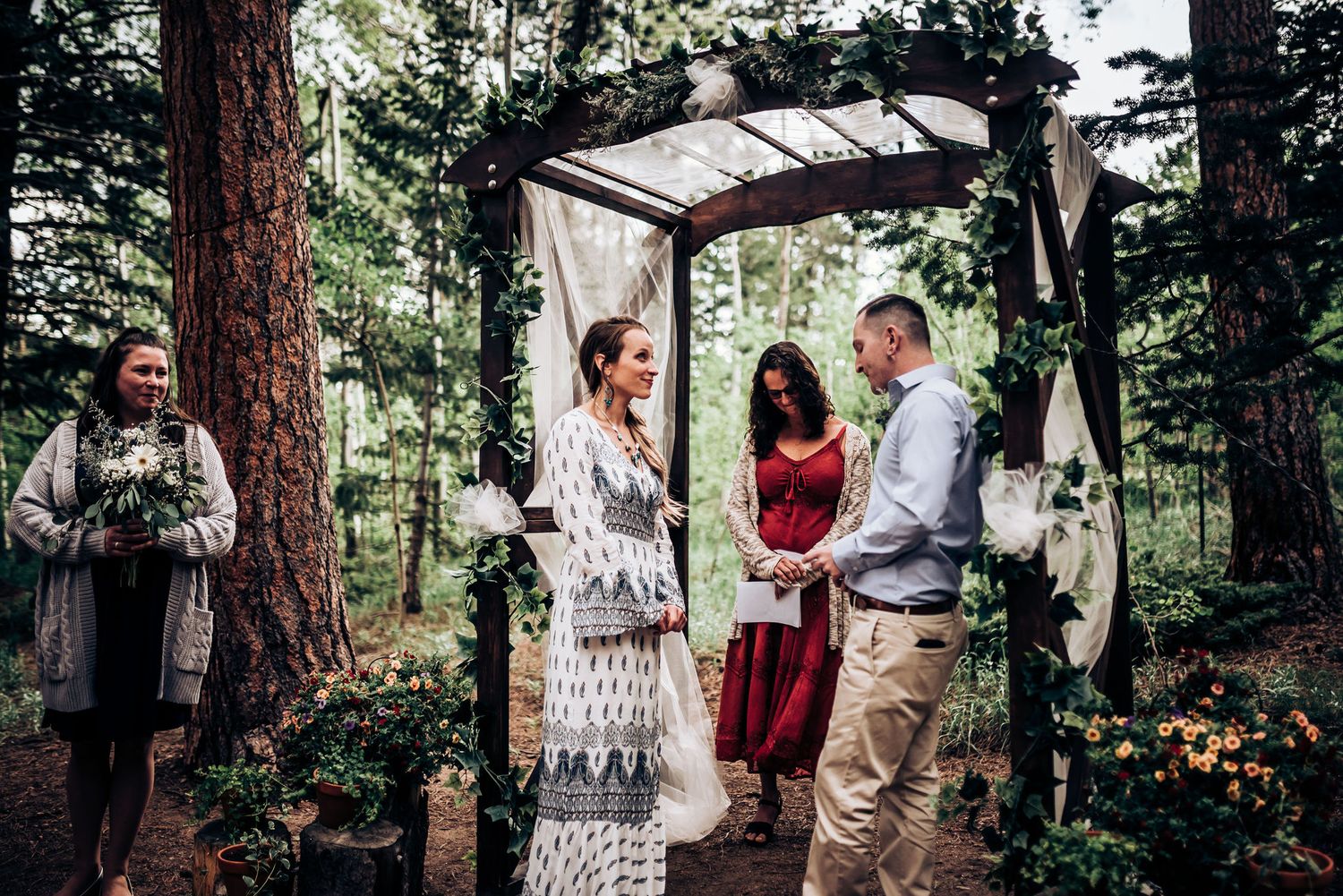 A wedding ceremony unfolds beneath a decorated arch in a serene forest setting.