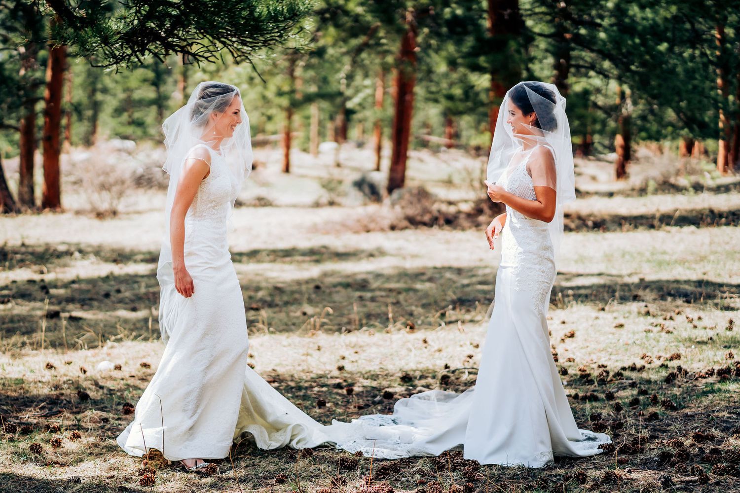 Two brides in fitted white wedding dresses stand together in a wooded outdoor setting.