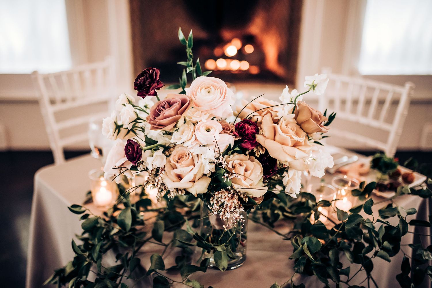 Romantic floral centerpiece with pink roses and greenery near a glowing fireplace.