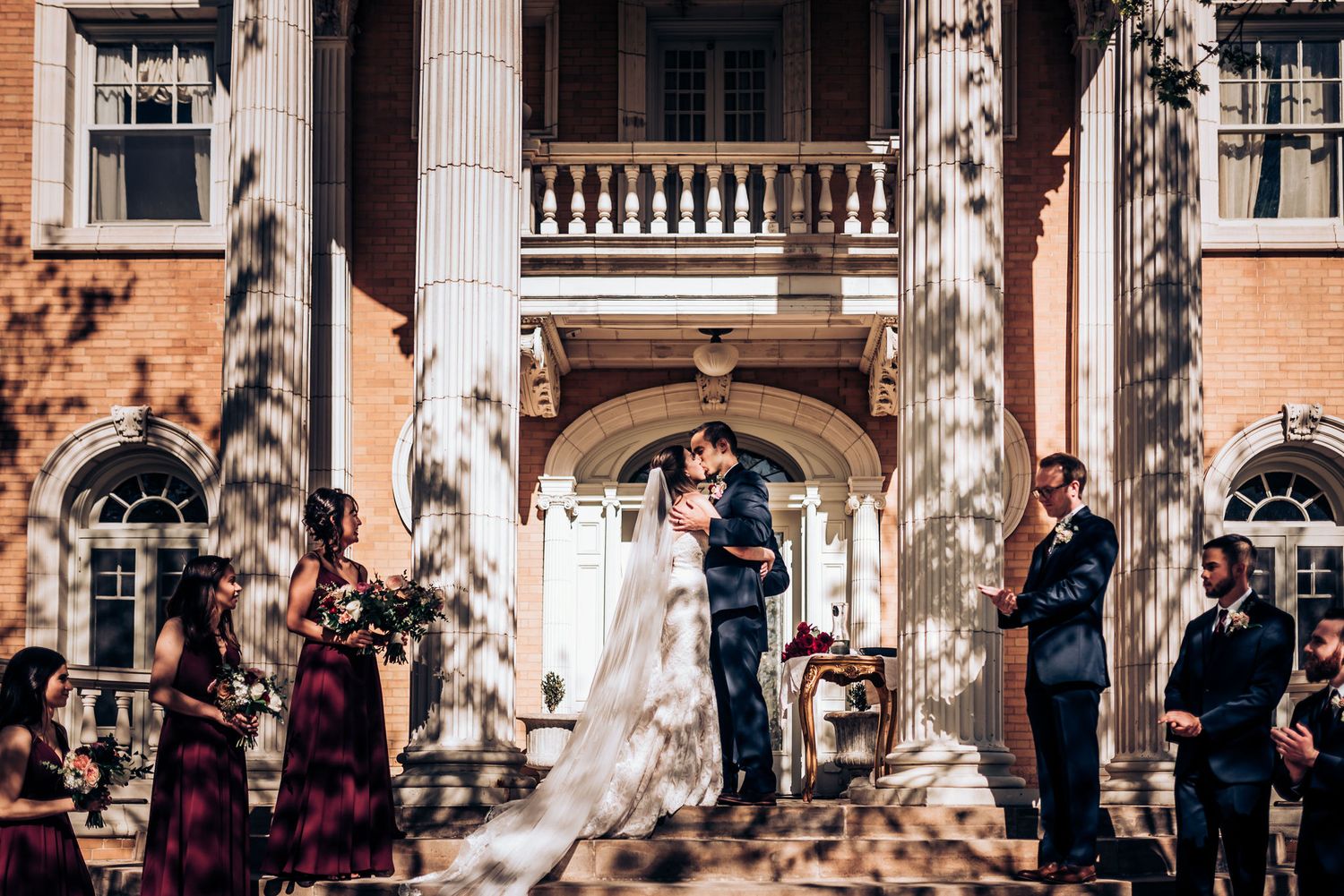 Wedding ceremony on sunlit steps of historic building with white columns and brick facade.