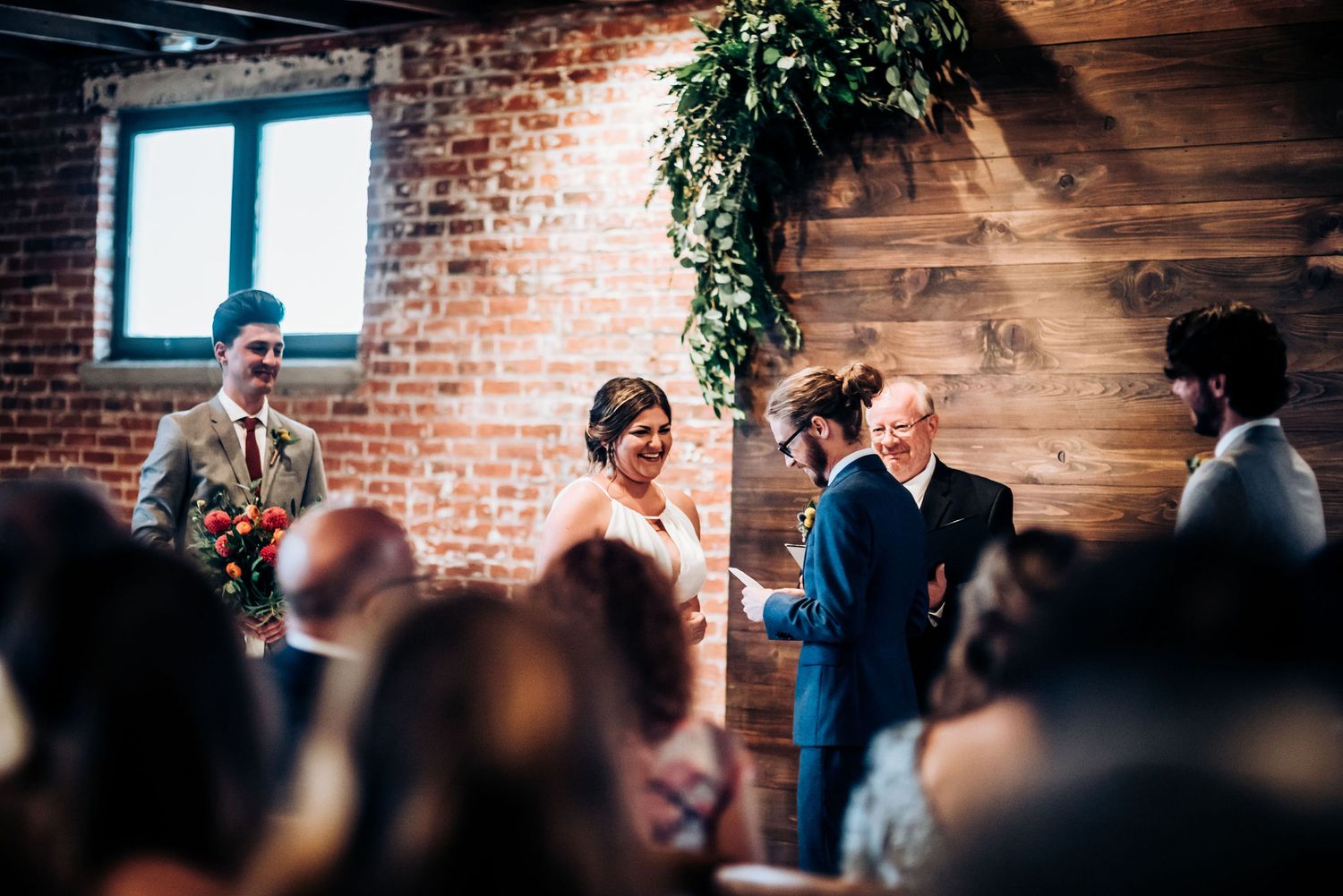 Wedding ceremony in progress at industrial-chic venue with brick walls and hanging greenery.