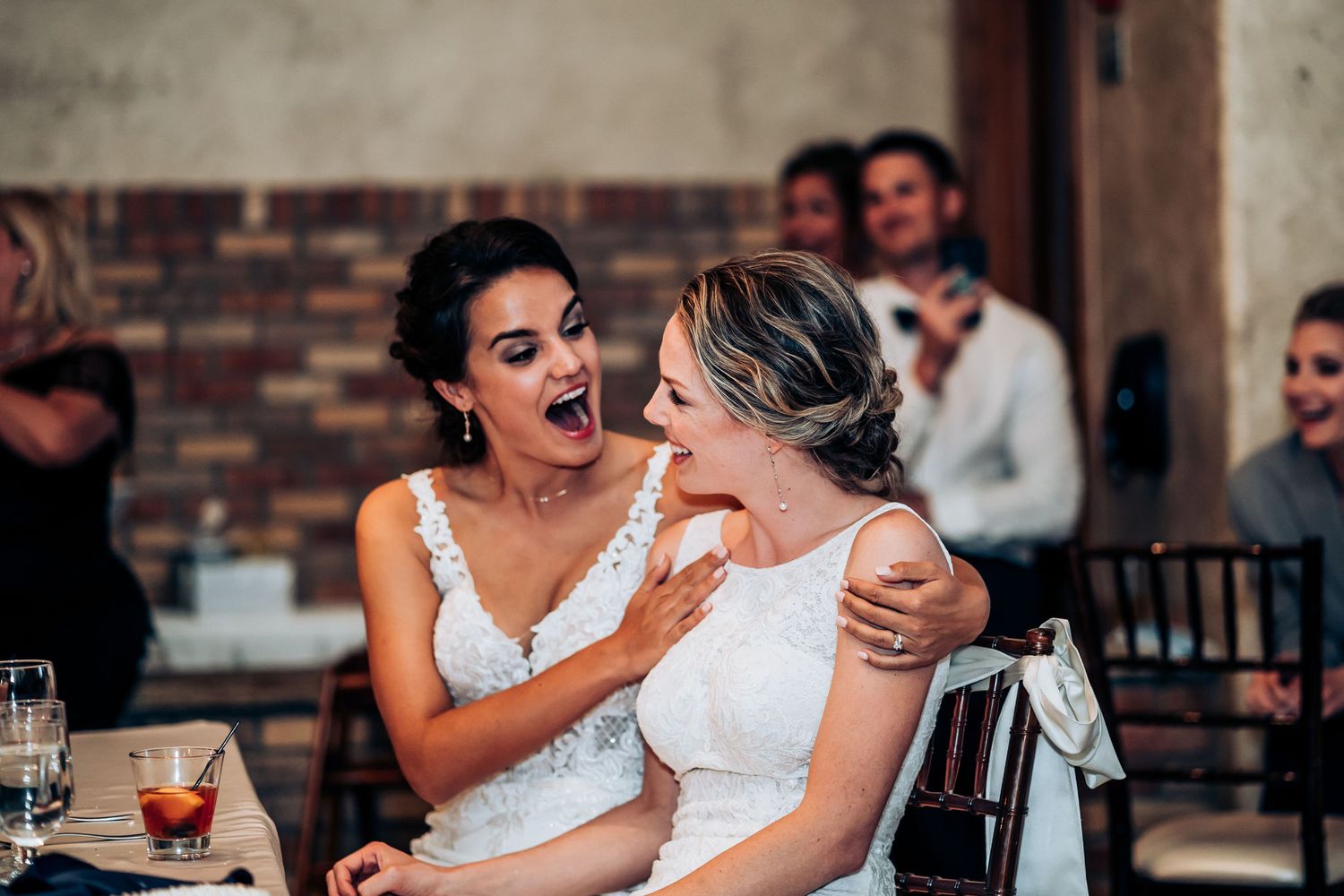 Two brides share an emotional candid moment while laughing together during wedding reception toasts.