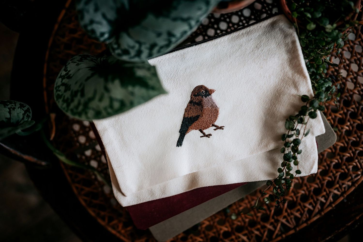 White fabric pouch with detailed bird embroidery surrounded by greenery.