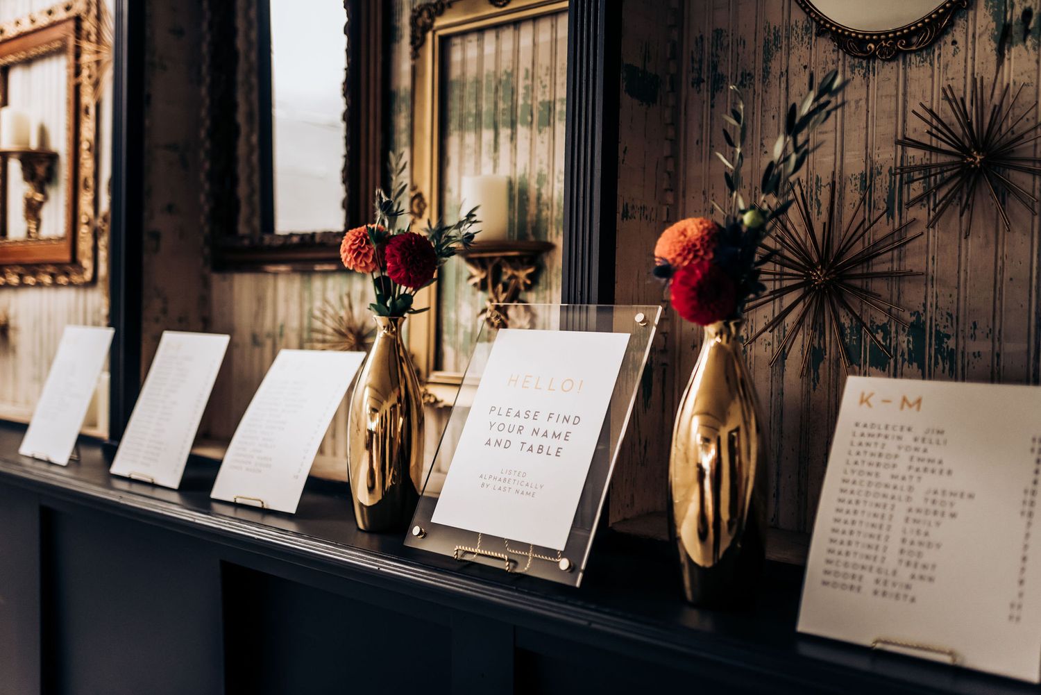 Wedding seating cards displayed on a wooden shelf with red roses in gold vases.