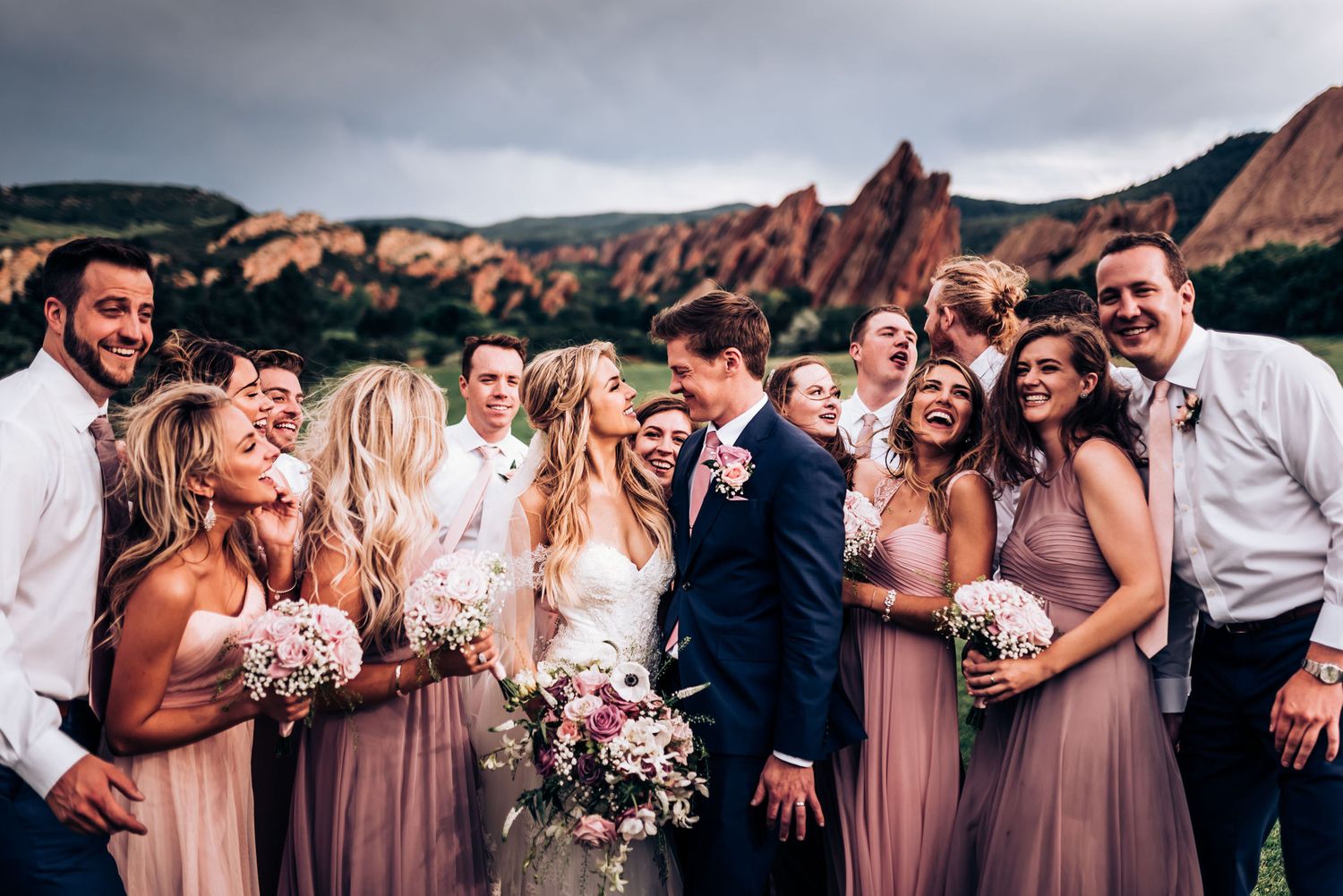 Wedding party laughs together during outdoor photo session with mountain backdrop.