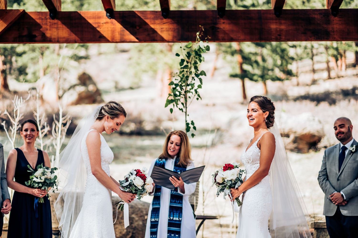 Wedding ceremony in progress under wooden pergola with bridal party in attendance.