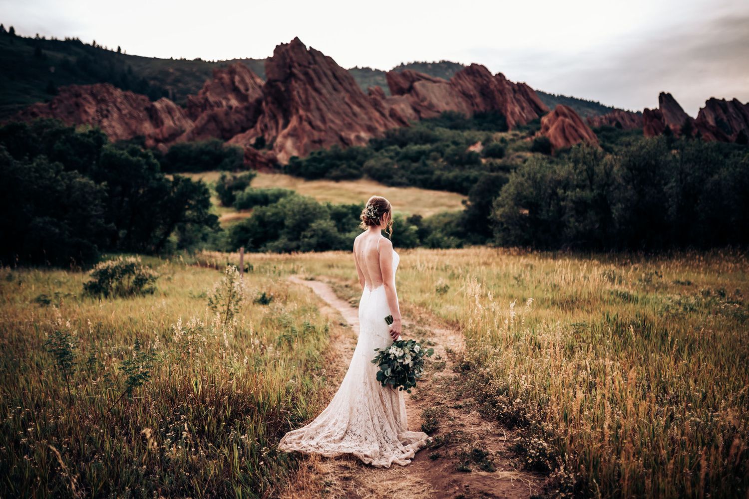 A bride in a flowing dress stands on a trail with dramatic red rock formations in the background.
