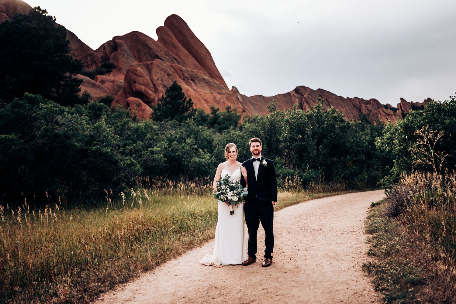 Newlyweds walking on trail with majestic red rock formations in background.