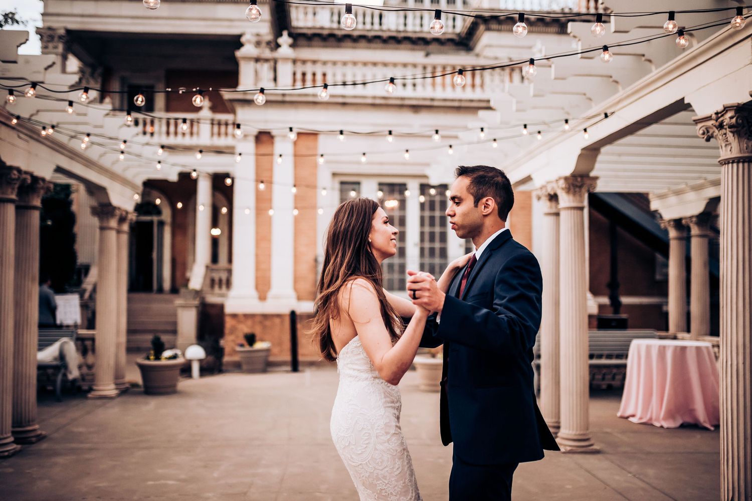 Couple shares first dance under string lights in elegant columned reception space.