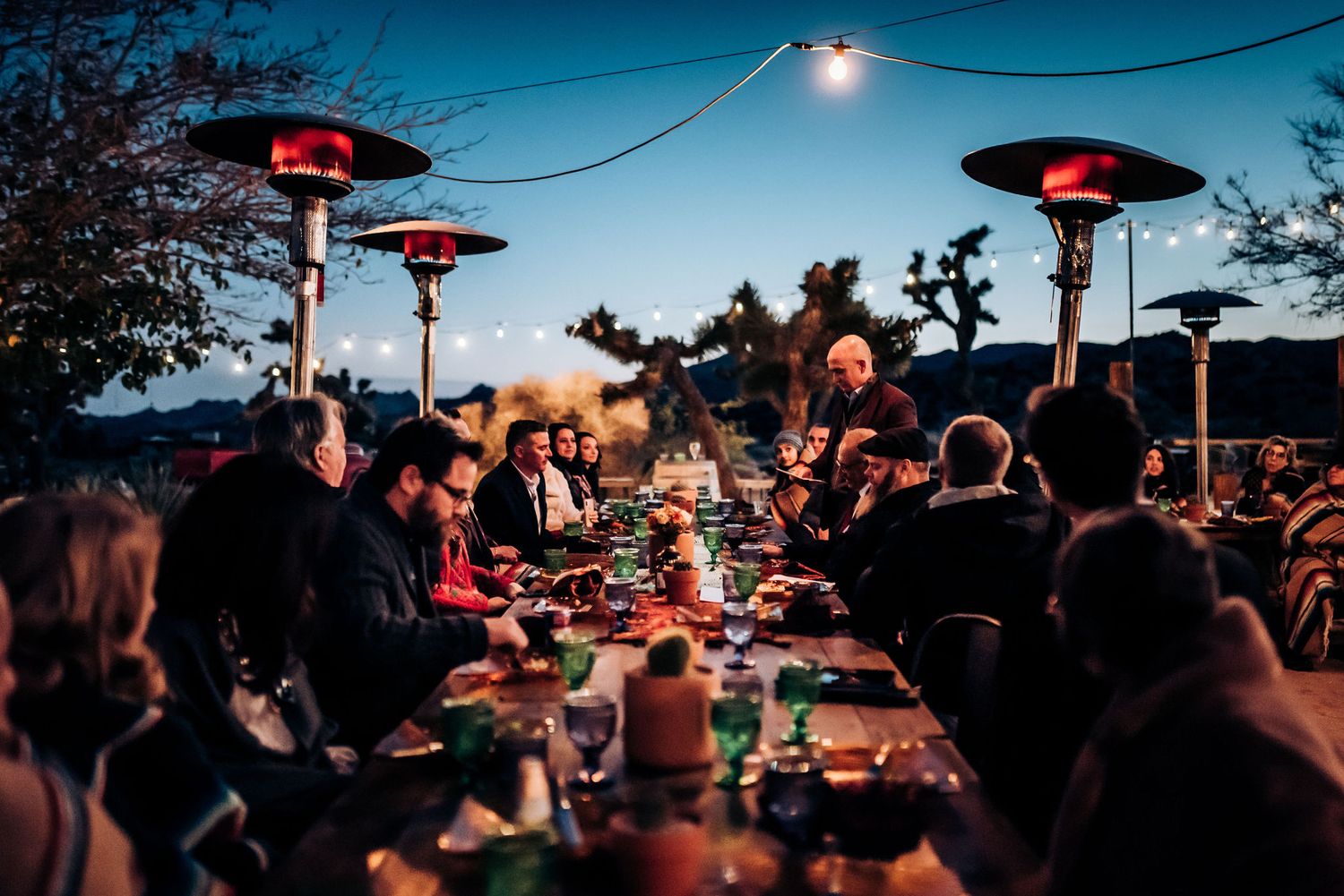 Friends enjoying dinner under warm heat lamps and string lights at a long outdoor table.