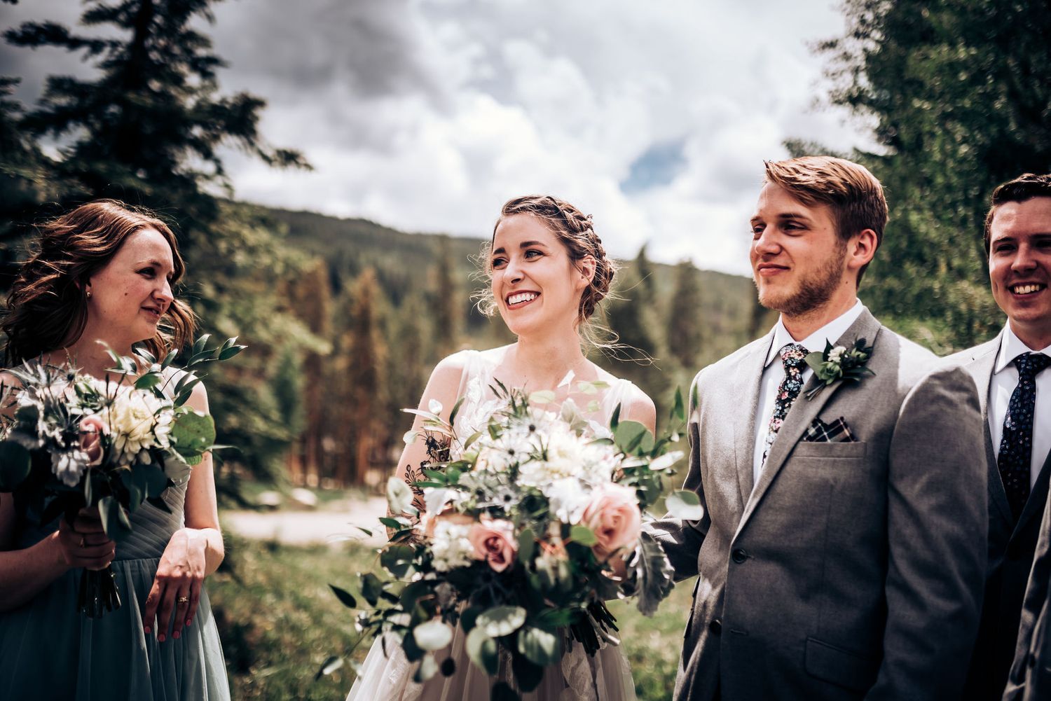Wedding party laughing together against mountain backdrop with rustic bouquets in forest setting.