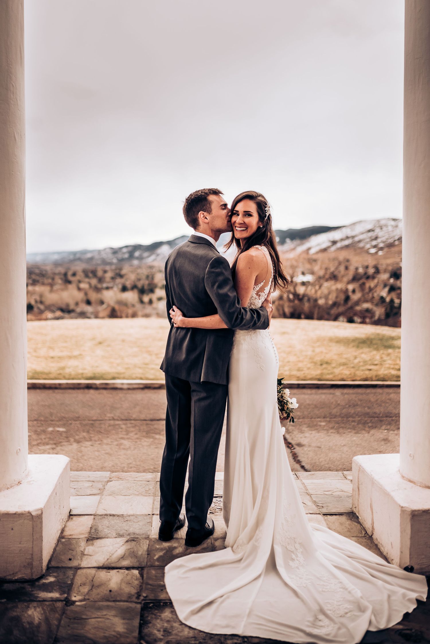 Romantic wedding photo of couple embracing on a columned terrace overlooking scenic mountain landscape.
