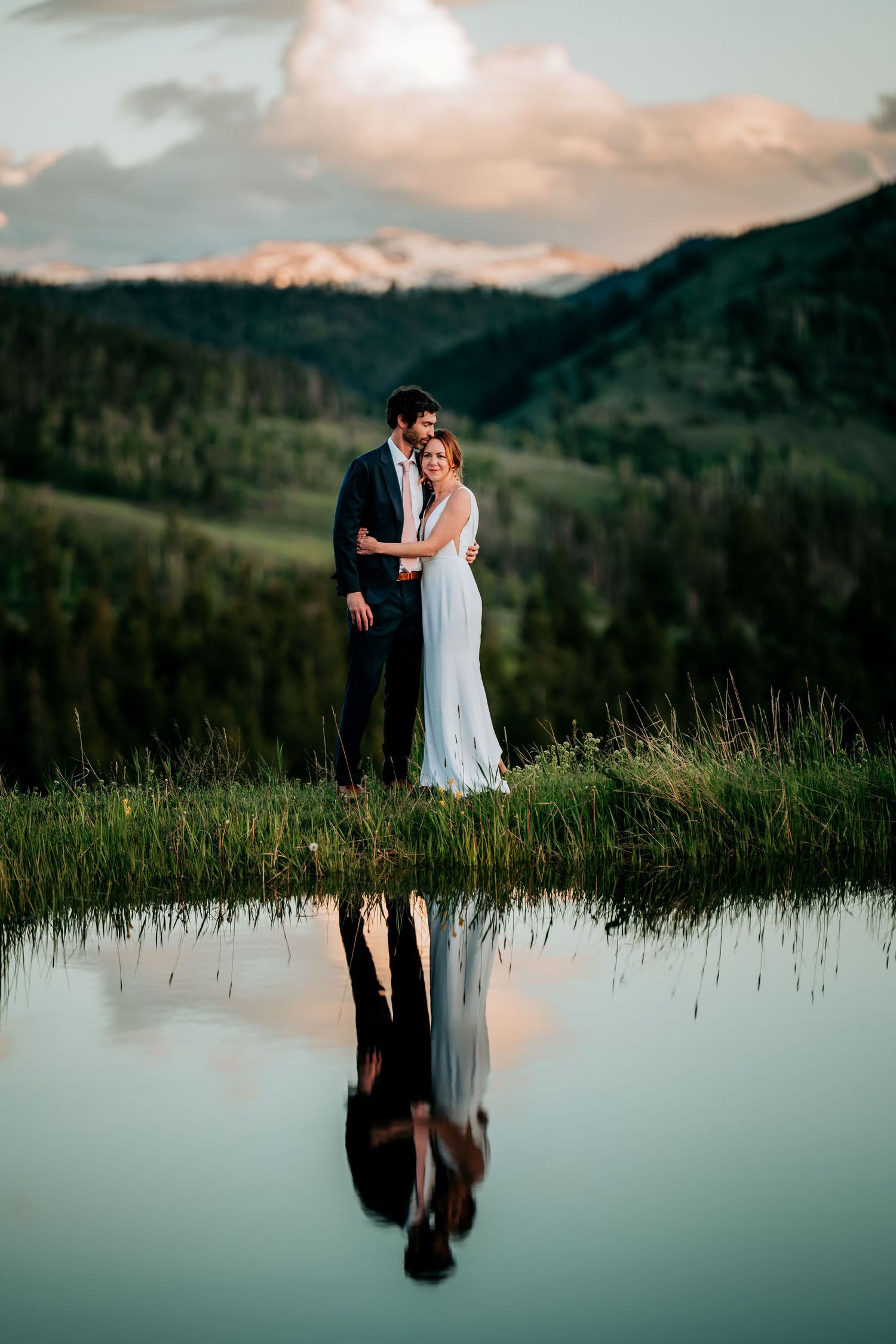 Romantic mountain wedding portrait with couple's reflection in calm lake at sunset.