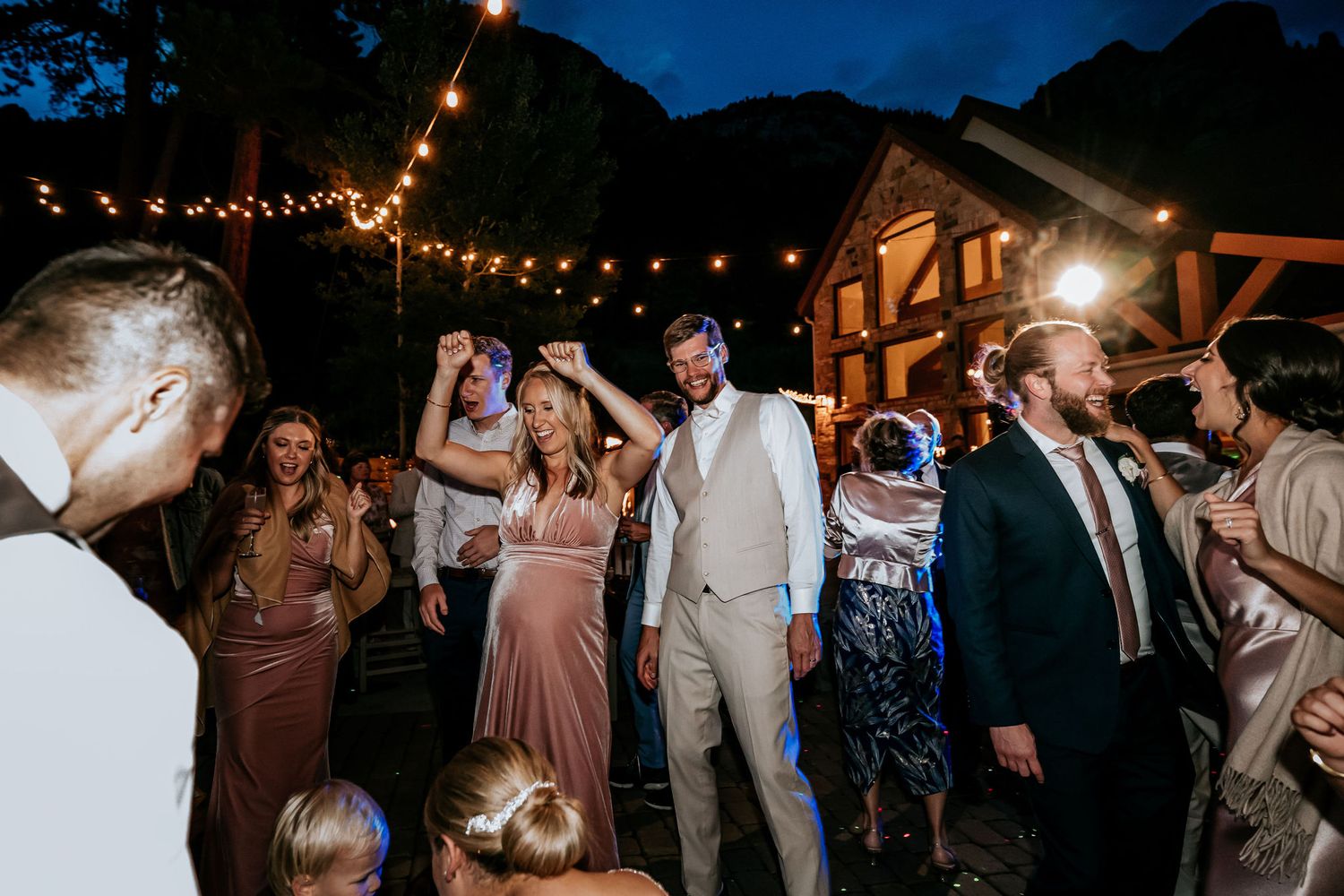 Guests celebrate on a dance floor beneath rustic string lights with mountains in the background.