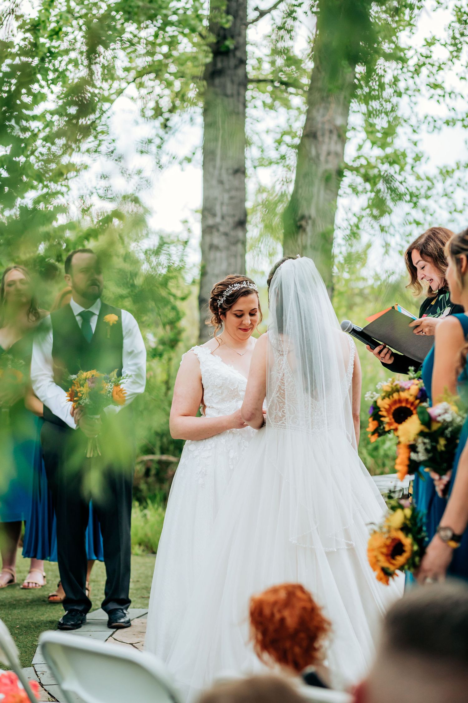 Outdoor wedding ceremony under tall trees with sunflowers in the bridal bouquet.
