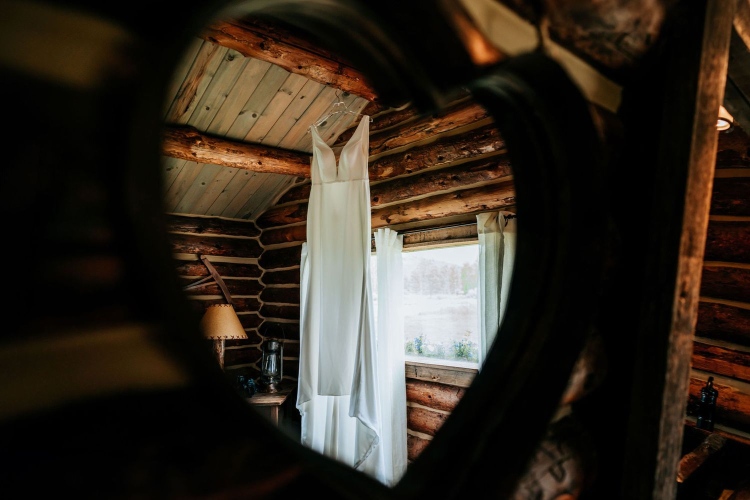 Wedding dress hanging in rustic log cabin interior viewed through mirror reflection.