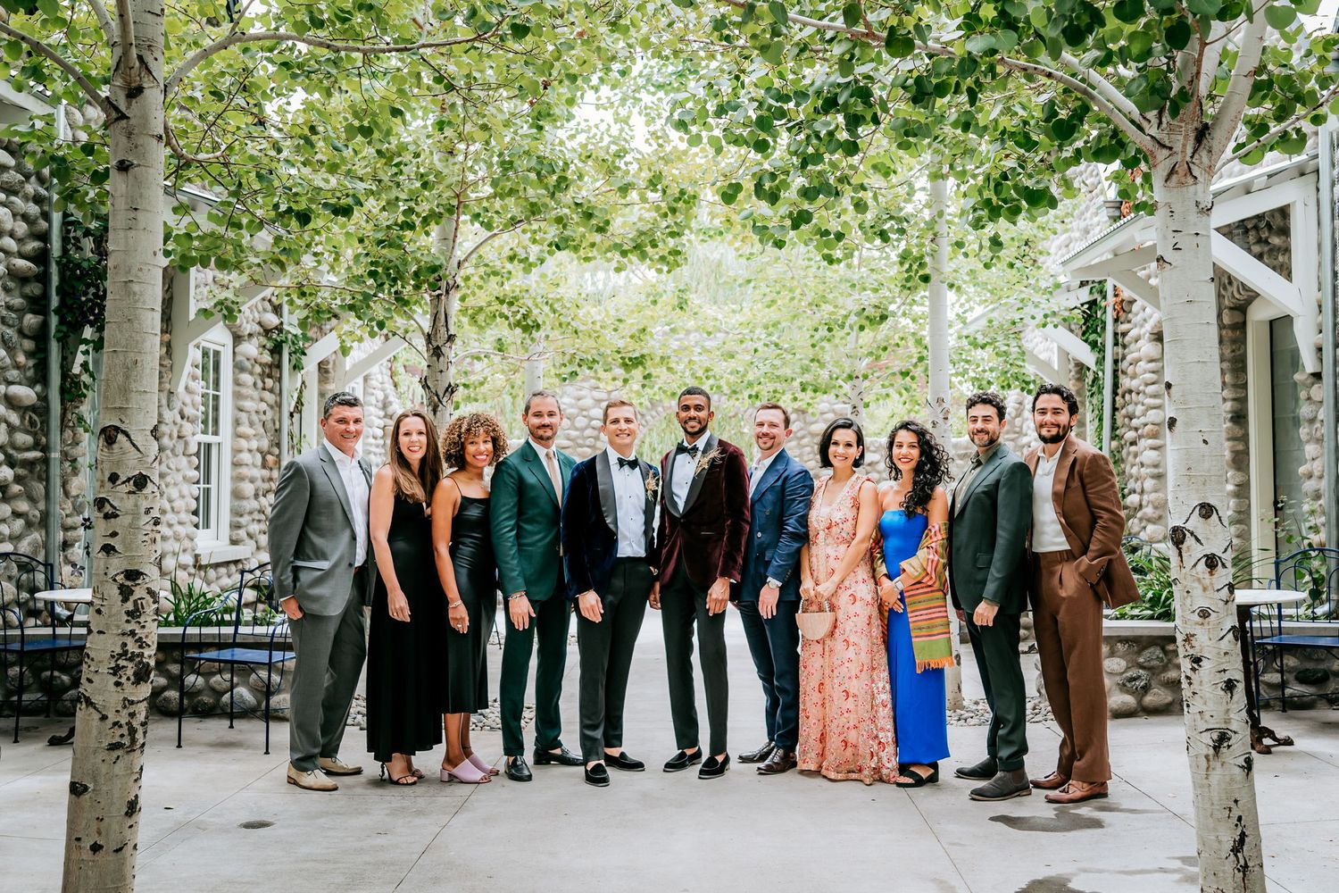 Group photo of formal-dressed guests standing together between stone pillars and trees in an outdoor setting.