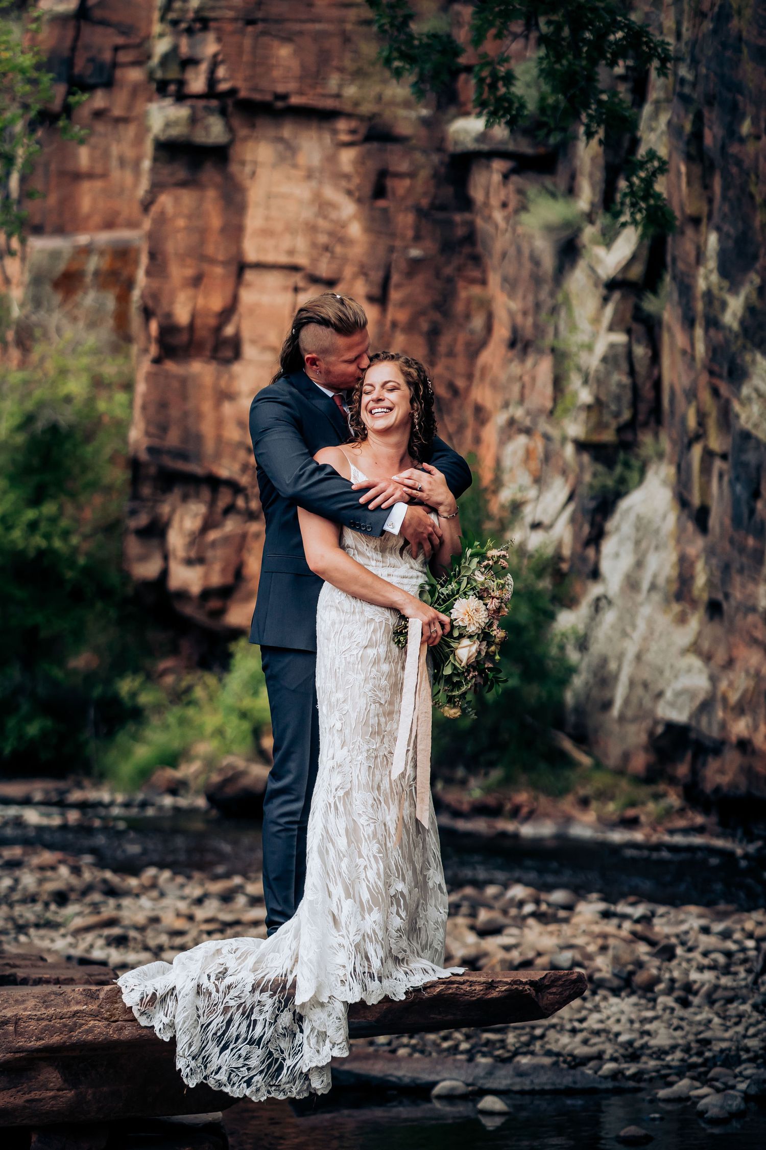 Romantic wedding portrait captured against dramatic red rock cliffs near water.