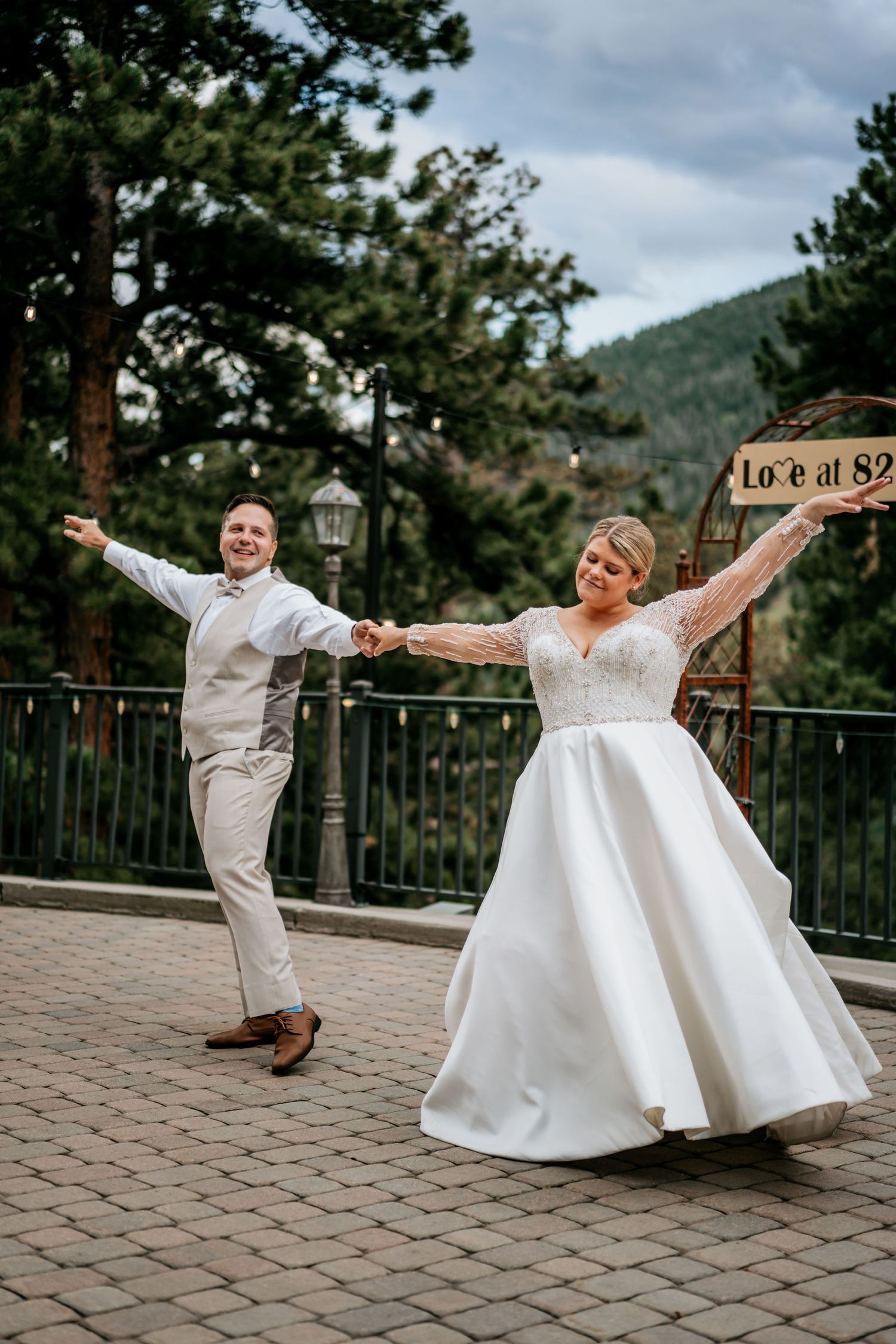 A joyful wedding couple dances on a patio with mountains and pine trees in the background.