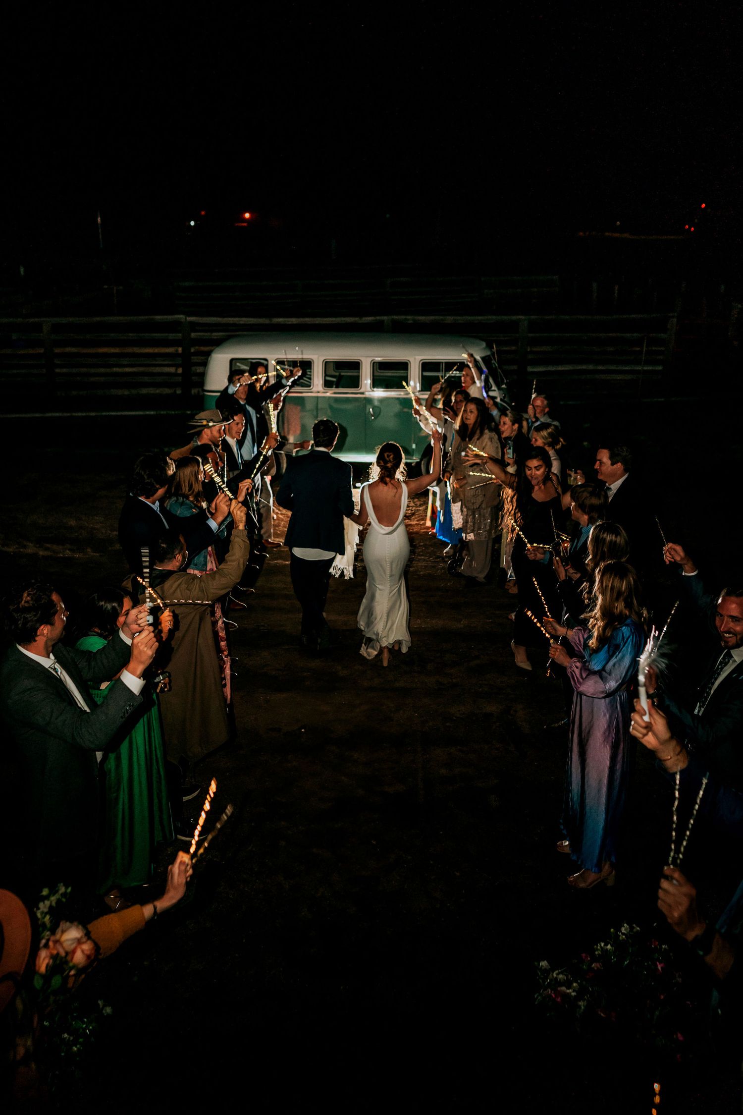 Wedding party exits through sparkler tunnel at night with vintage VW bus in background.