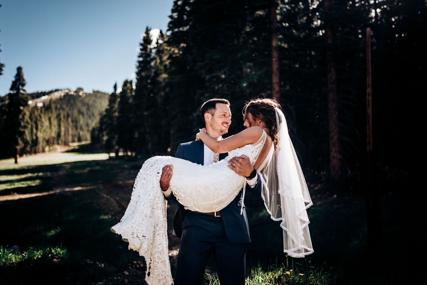 Wedding couple shares romantic moment in mountain forest setting as groom carries bride in white lace dress.