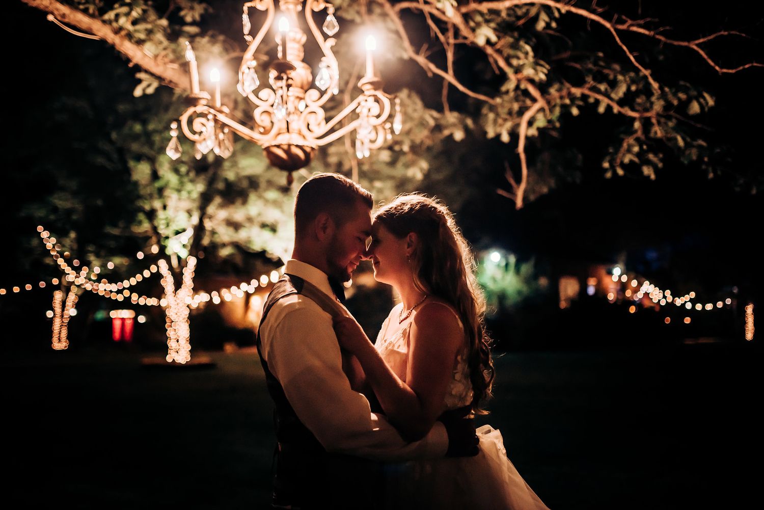 A romantic evening dance under a crystal chandelier and string lights in an outdoor garden setting.