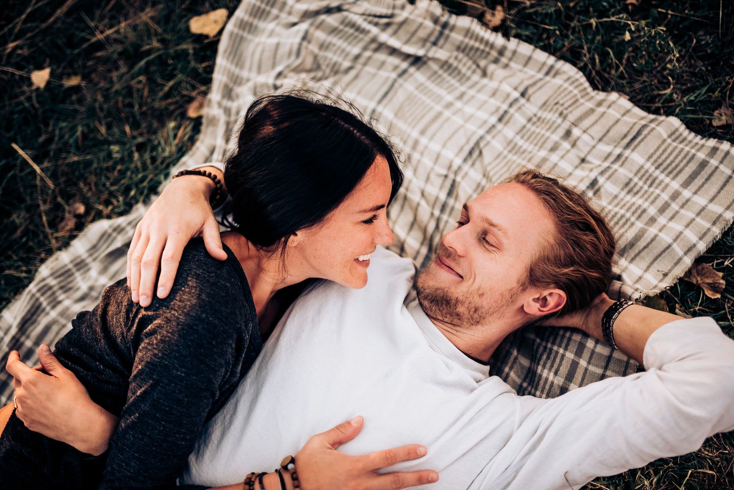 A couple lies on plaid blanket sharing intimate moment outdoors.