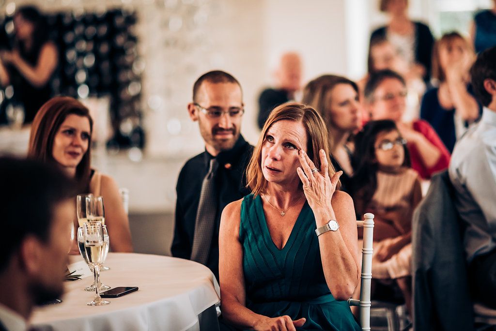 Wedding guests react emotionally during speeches at an indoor reception with decorative lighting.