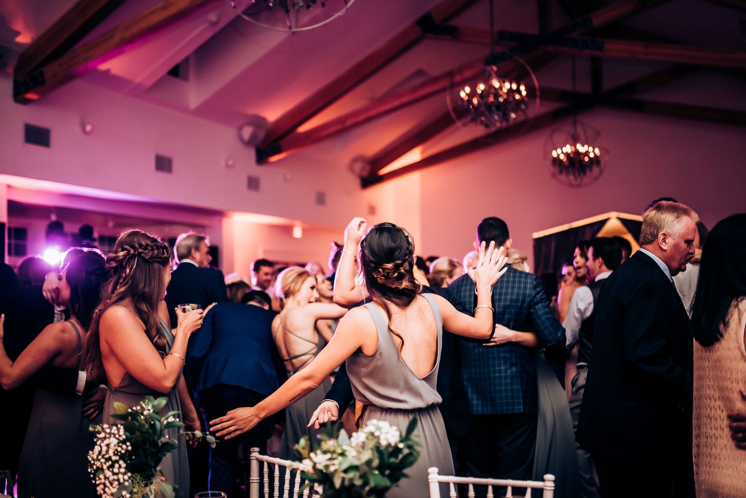 Wedding guests dance and celebrate on a pink-lit dance floor under exposed wooden beams.