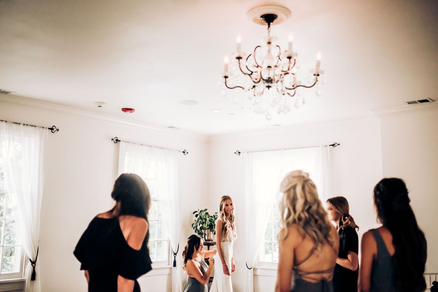 Wedding party gathers around bride in bright white room with chandelier.