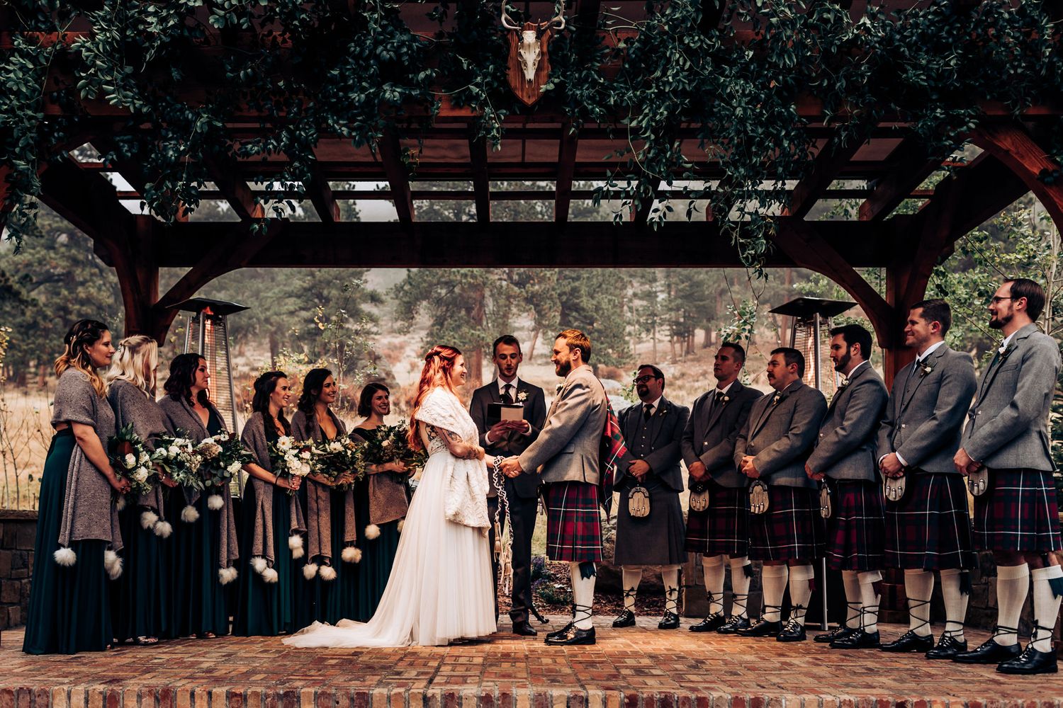 Wedding party gathers under pergola with bridesmaids in gray and groomsmen in traditional Scottish kilts during ceremony.