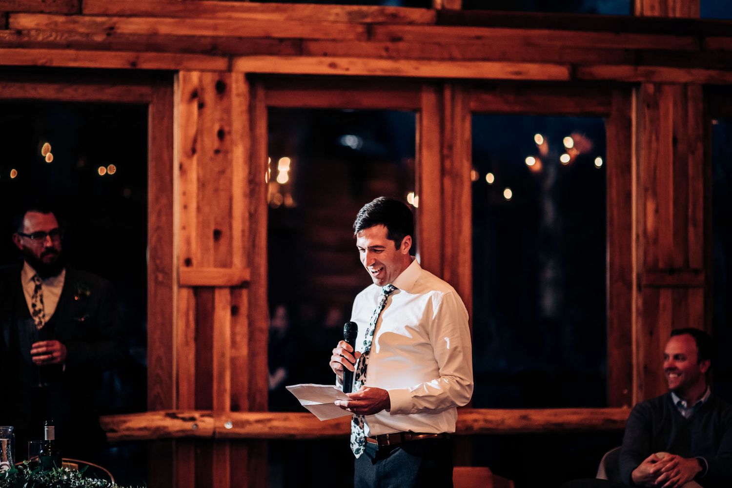 Speaker giving toast at wedding reception in front of wooden wall and windows.