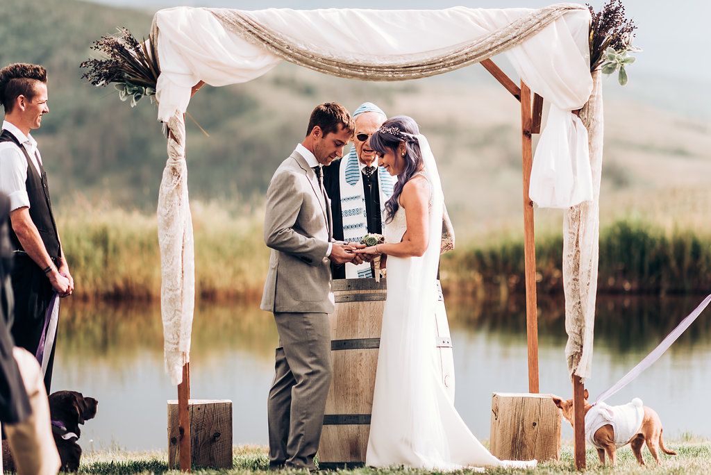 A rustic outdoor wedding ceremony under a draped fabric arch with a wooden barrel and serene lake backdrop.