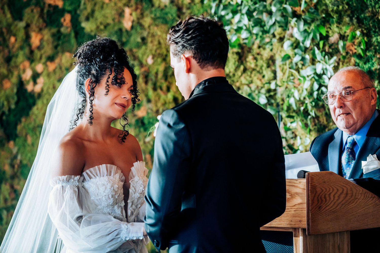 Wedding ceremony taking place in front of lush green foliage wall with hanging lights.