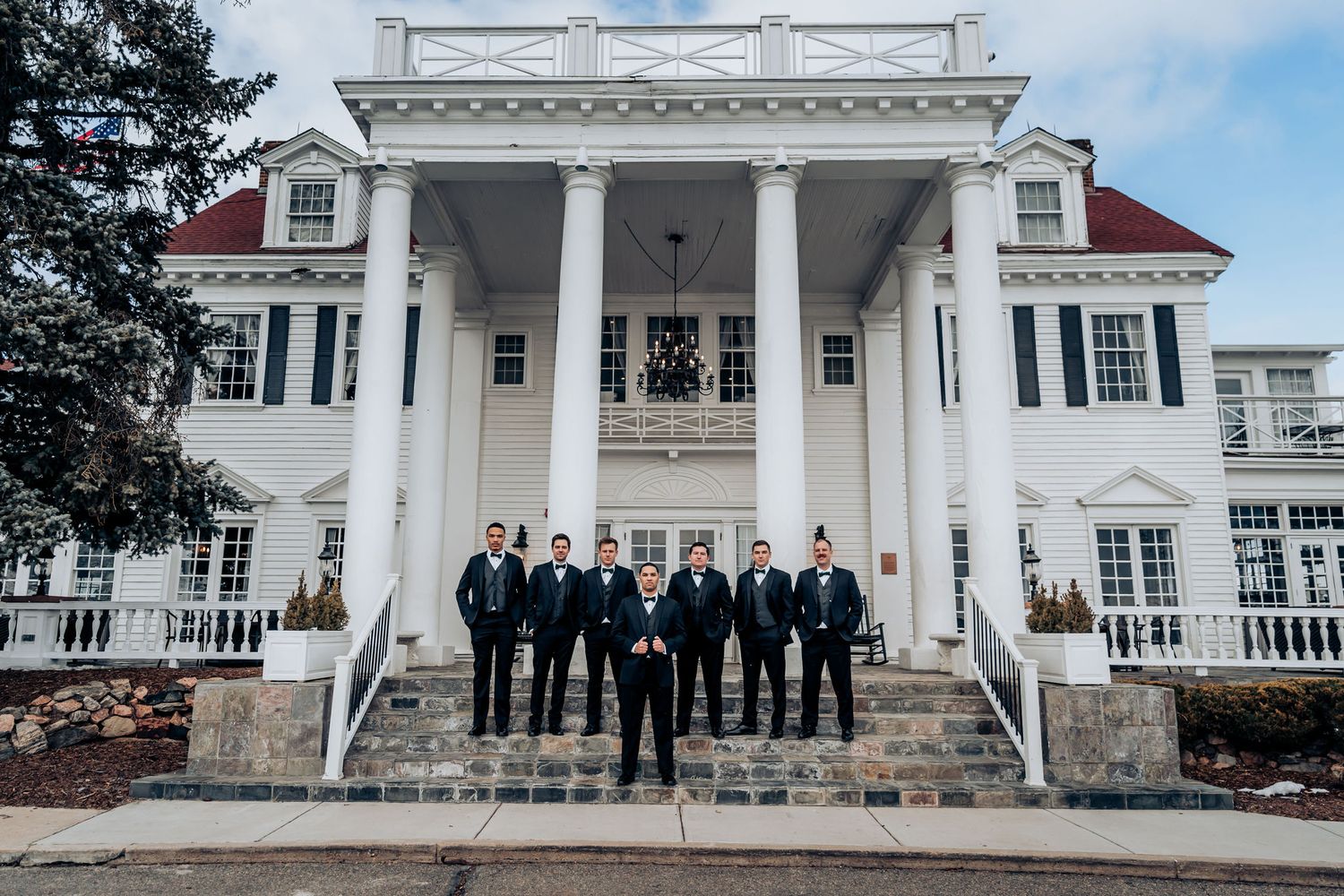 Wedding party stands in front of elegant white mansion with grand columns and red roof.
