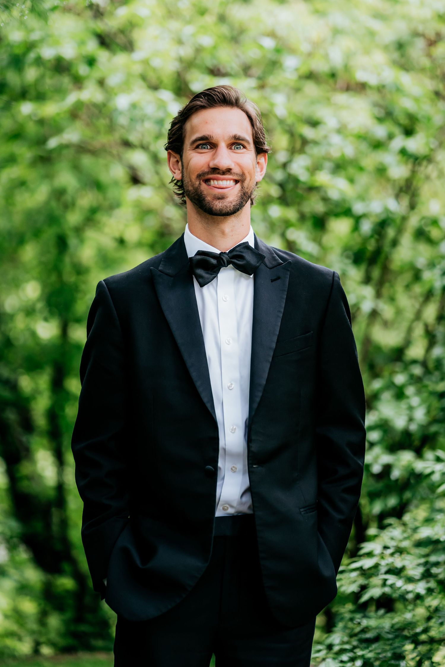 Groom in classic black tuxedo and bowtie smiling against green foliage background.