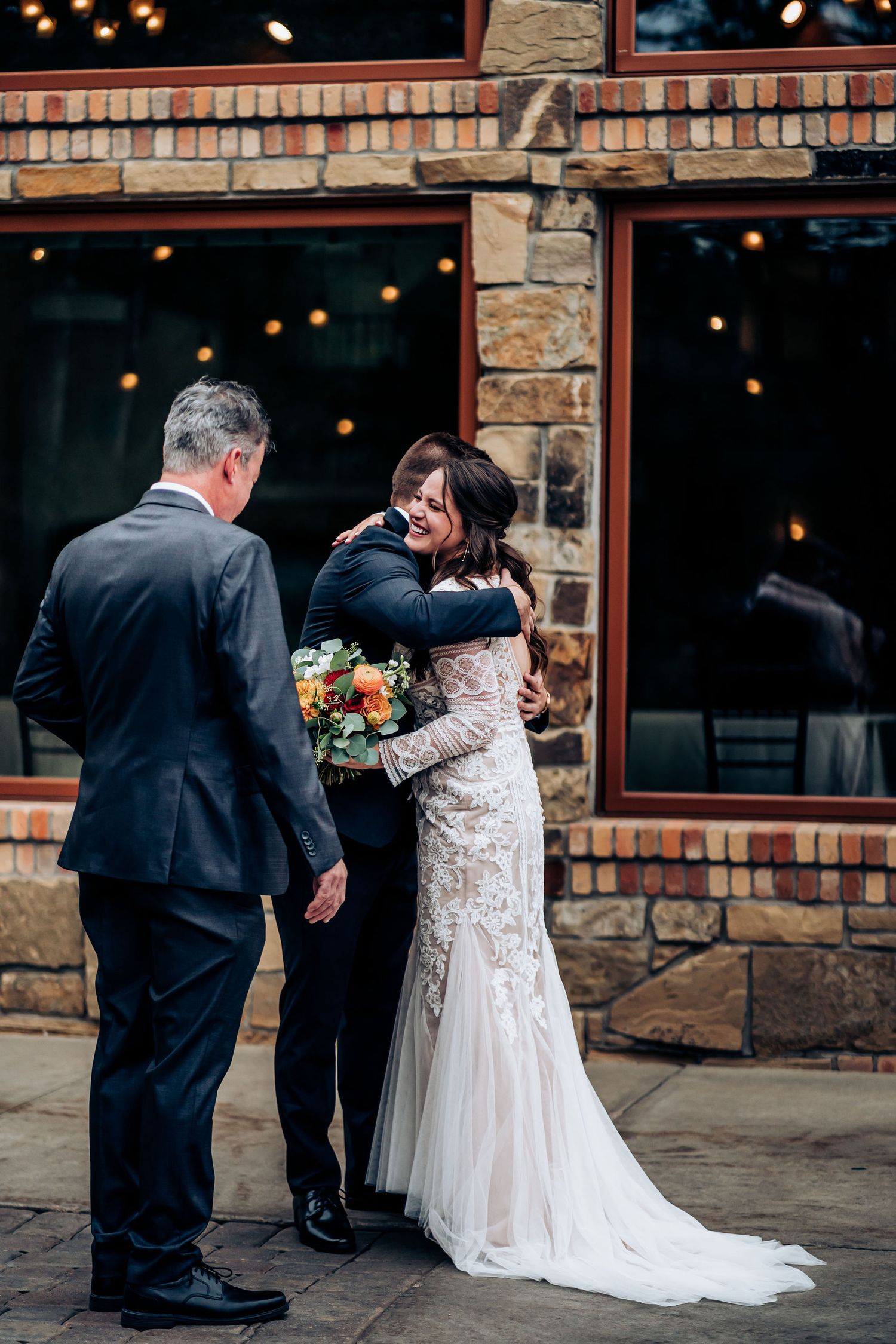 Emotional wedding moment shared between couple and officiant outside rustic stone and brick venue with string lights.