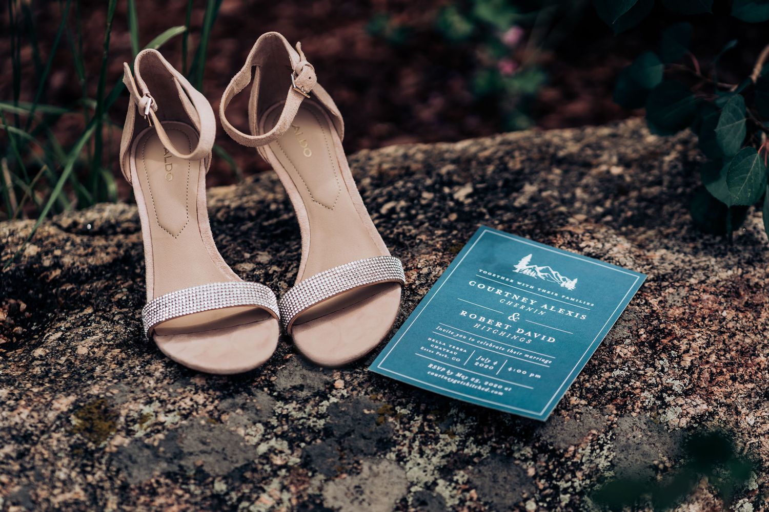 Nude wedding heels and a teal invitation card placed on a stone surface.