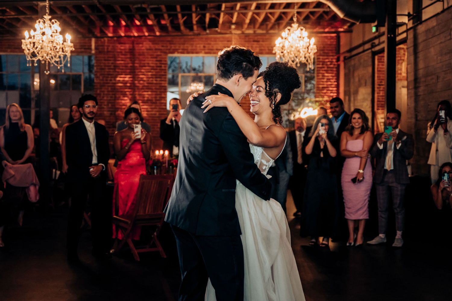 First dance at wedding reception in romantic venue with exposed brick walls and chandelier lighting.