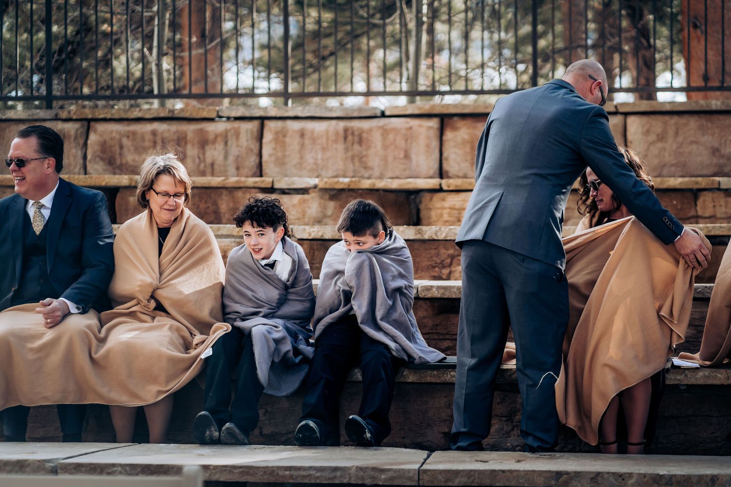 Wedding guests bundled in coats sit on wooden bench during outdoor winter ceremony.