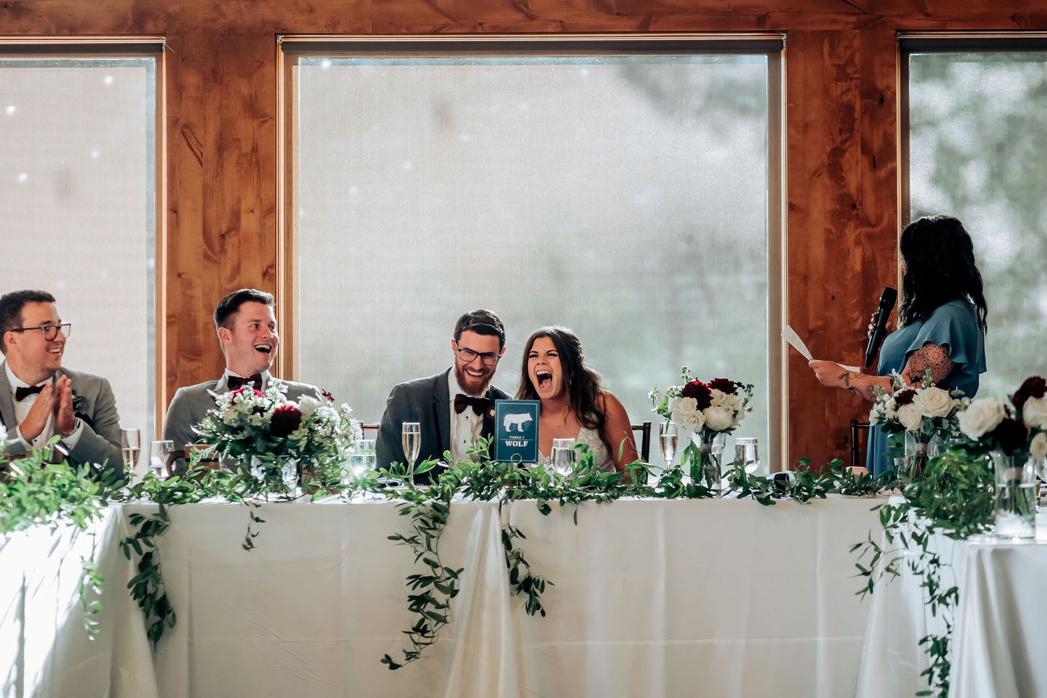 Wedding party shares laughs at head table decorated with flowing greenery garland.