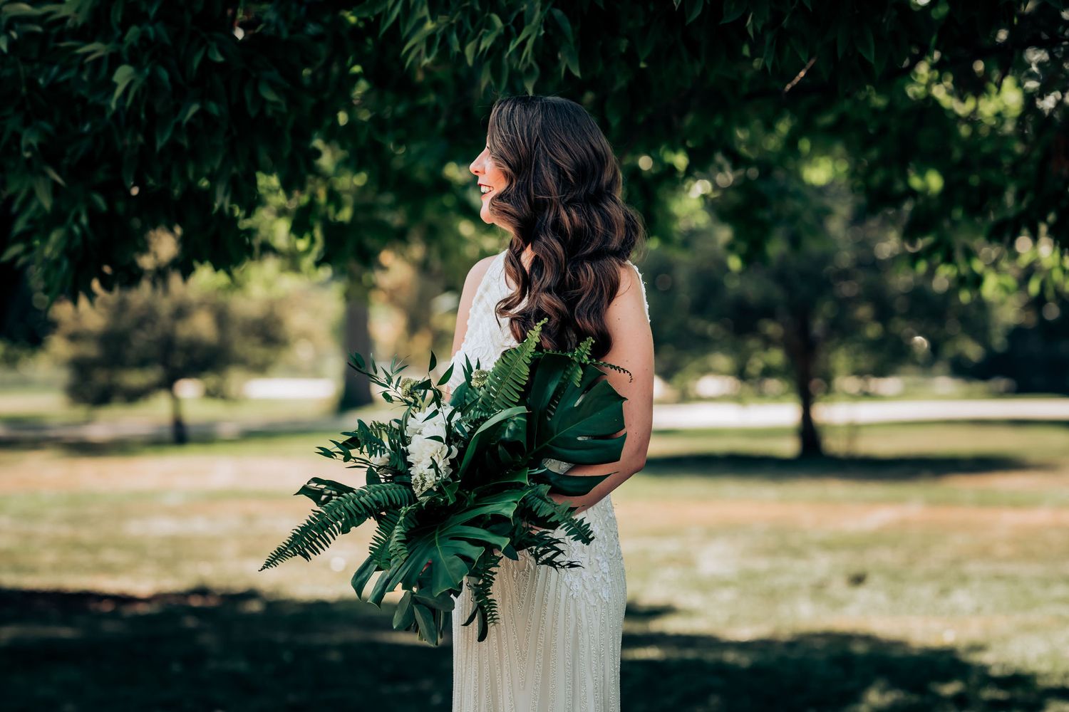 A side-view bridal portrait with flowing white dress and dramatic tropical bouquet in a park setting.