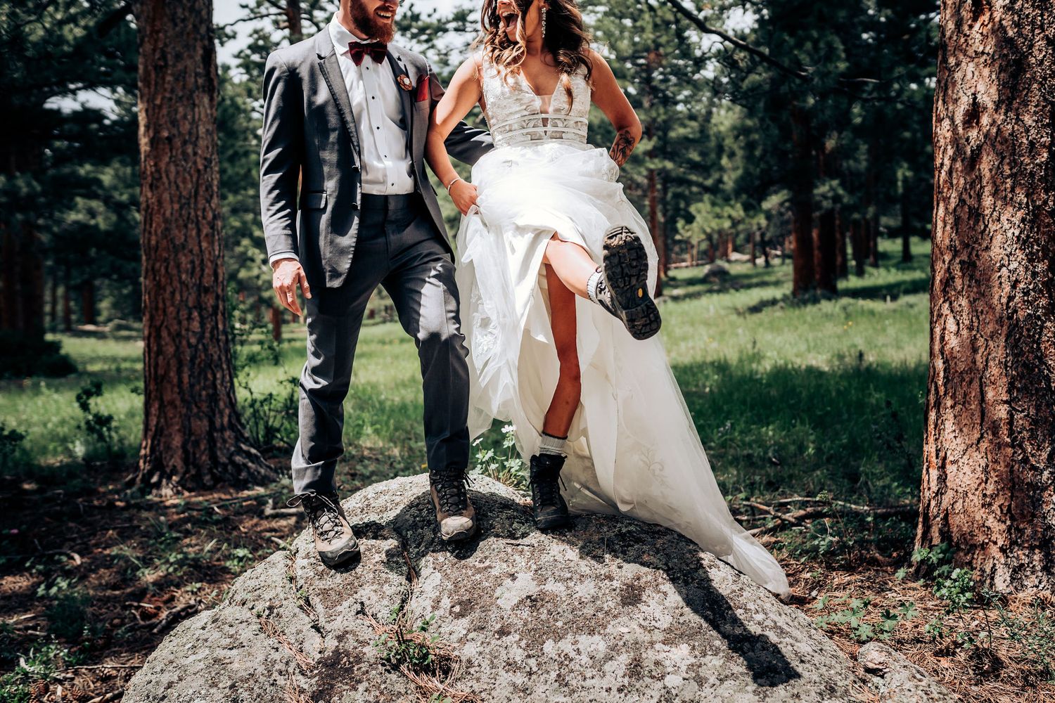 Wedding couple showing off their hiking boots while standing on a large boulder in a forest setting.