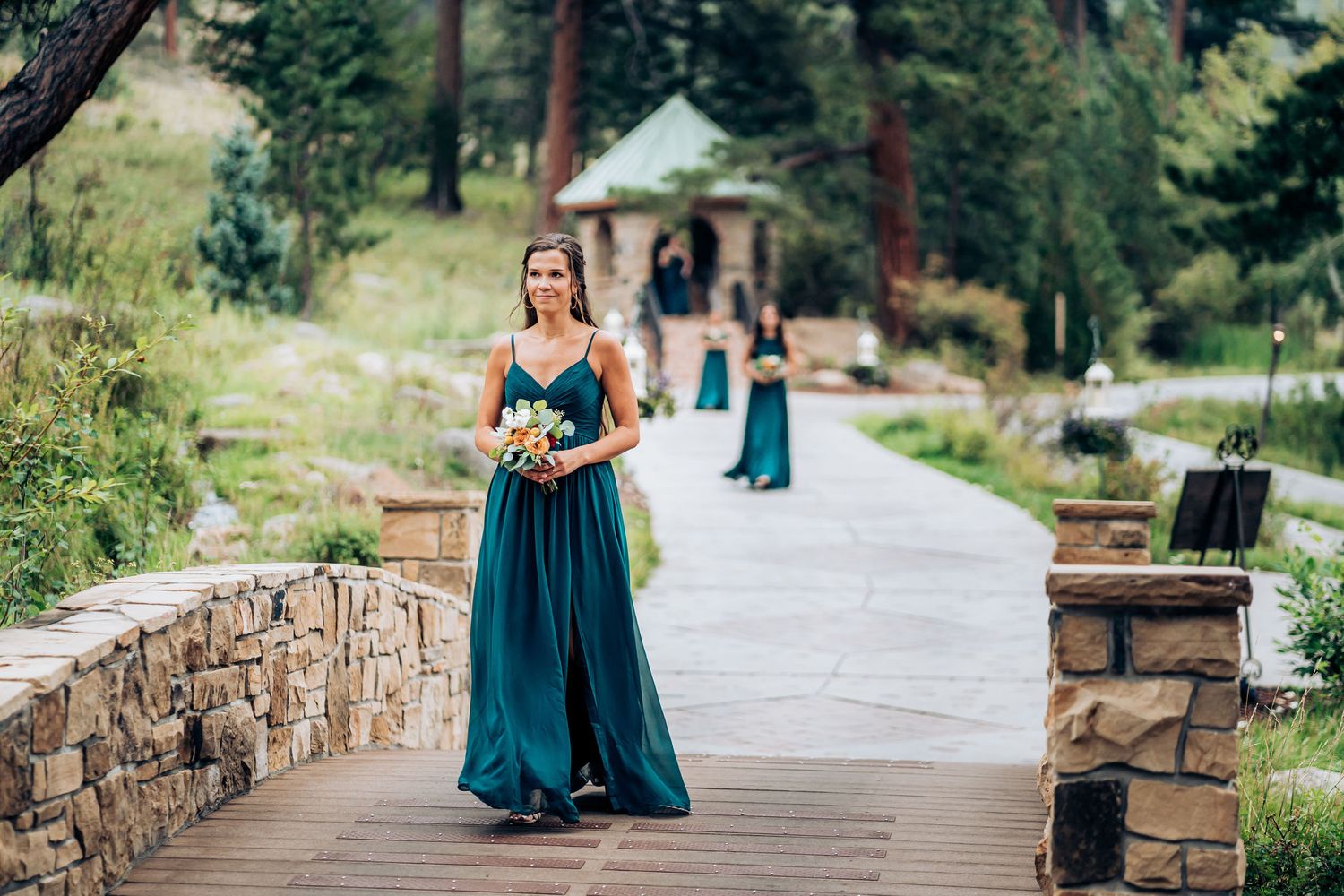 Bridesmaid in teal gown walking down outdoor pathway holding bouquet.