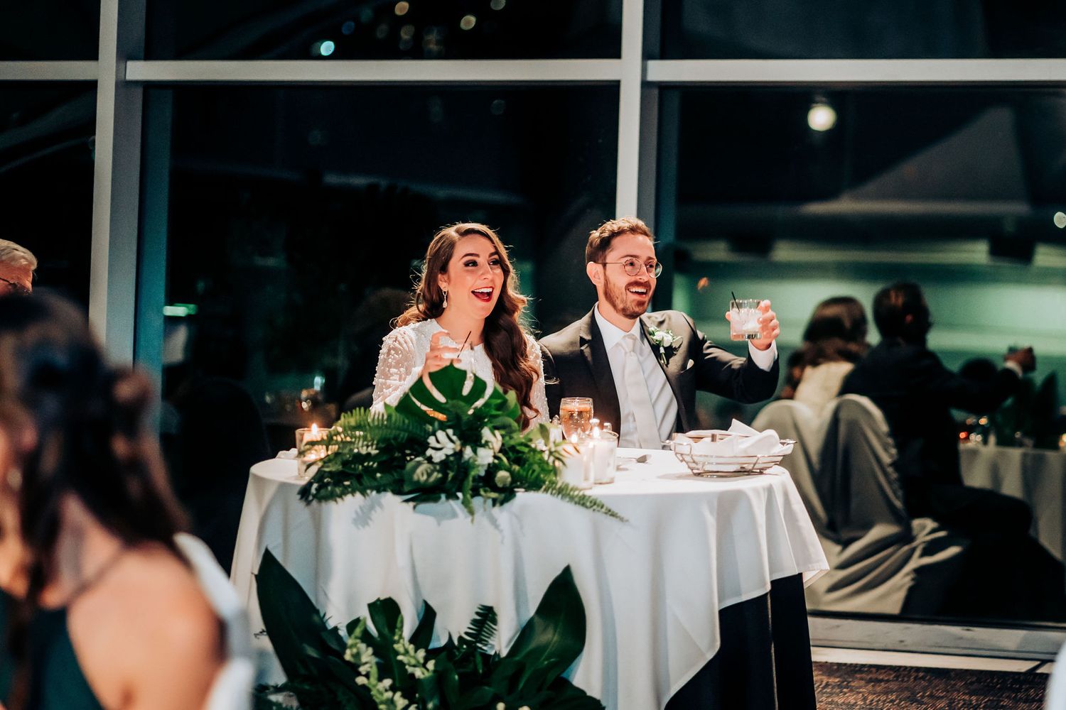 A couple shares a laughter-filled moment at their reception table adorned with greenery.