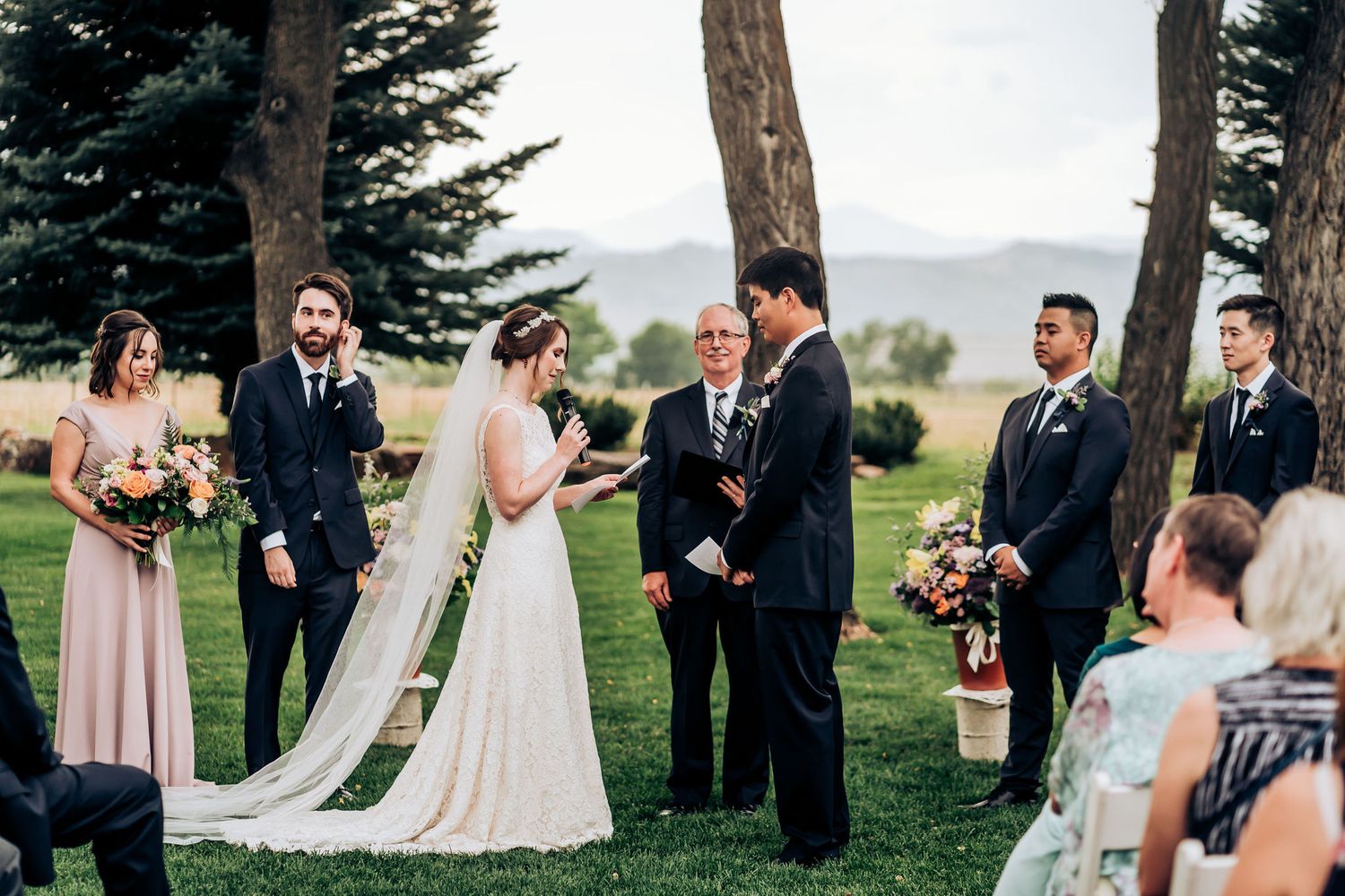 Wedding party gathered on lush grass with evergreen trees and mountains in the background.