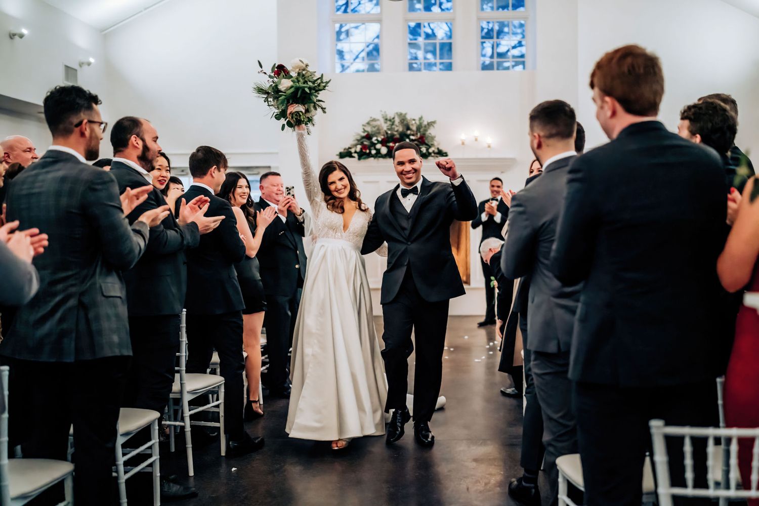 The newlyweds walk down the aisle while guests cheer and take photos in a modern chapel setting.